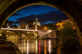 St. Peter's Basilica in Vatican City viewed through the arch of a bridge over the Tiber River in Rome, Italy, featuring illuminated architecture, reflections on the water, and a deep blue twilight sky.