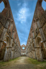 Looking through the roofless Gothic interior of the Abbey of San Galgano in Tuscany, Italy, featuring soaring stone arches, symmetrical architecture, and an open sky above.