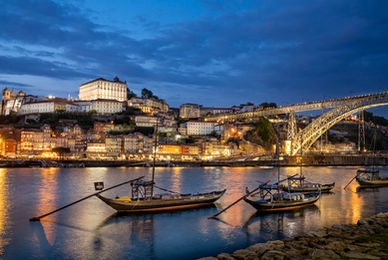 The Dom Luís I Bridge spanning the Douro River in Porto, Portugal, at twilight, featuring traditional rabelo boats, the illuminated Ribeira waterfront, historic hillside architecture, and reflections on the water.