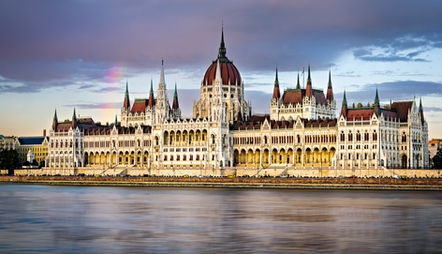 The Hungarian Parliament Building in Budapest, Hungary, viewed from across the Danube River, featuring Neo-Gothic architecture, soaring spires, the iconic central dome, and reflections on the water beneath a dramatic sky.