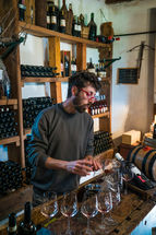 A sommelier preparing a wine tasting at a winery in Tuscany, Italy, with wine glasses, oak barrels, and shelves of wine bottles in a traditional tasting room.