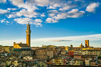 Panoramic view of Siena, Tuscany, Italy, featuring the Torre del Mangia, medieval buildings, terracotta rooftops, and a blue sky filled with scattered white clouds.
