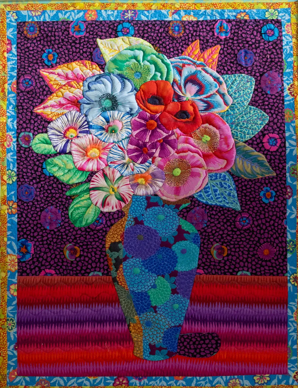 A vibrant textile art piece by Anna Wright featuring a blue patterned vase filled with "fussy cut" 3D flowers against a dark purple patterned background.
