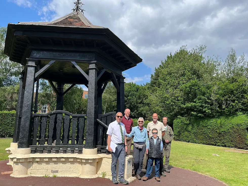 A group of six park professionals and volunteers standing in front of a traditional black wooden bandstand at John Innes Park.