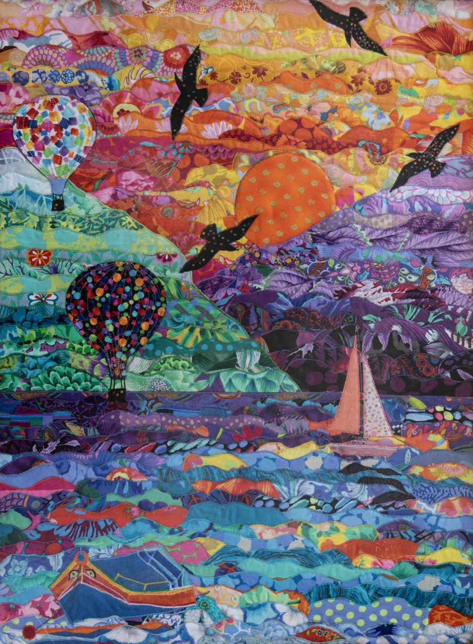 A large-scale textile landscape by Anna Wright with two hot air balloons, a sailing boat, and a large orange sun over a textured purple and blue sea.