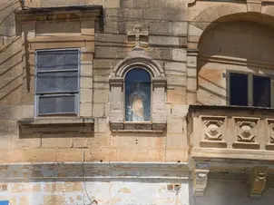 View of the Niche of the Sacred Heart of Jesus on a weathered stone building facade, situated between a window and a stone balcony.