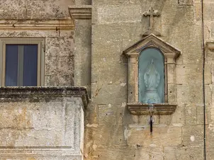 Side view of the Niche Madonna of Graces on a weathered stone building, showing the statue within a glass-covered recess, alongside a window and balcony.