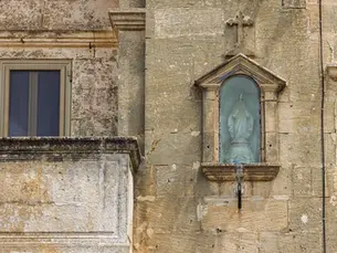 Side view of the Niche Madonna of Graces on a weathered stone building, showing the statue within a glass-covered recess, alongside a window and balcony.