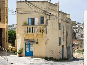 A full view of a light yellow building in Rabat, Gozo, with blue windows and doors, and a balcony.