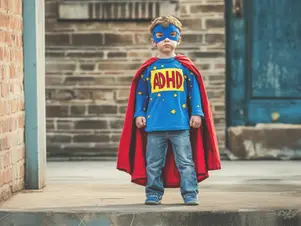 A young child dressed as a superhero with a red cape and a blue shirt featuring "ADHD" as an emblem, standing confidently in front of a brick building.