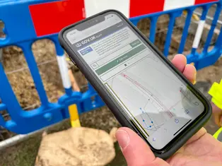 A construction worker holding a smartphone displaying the NUAR digital map interface at a work site