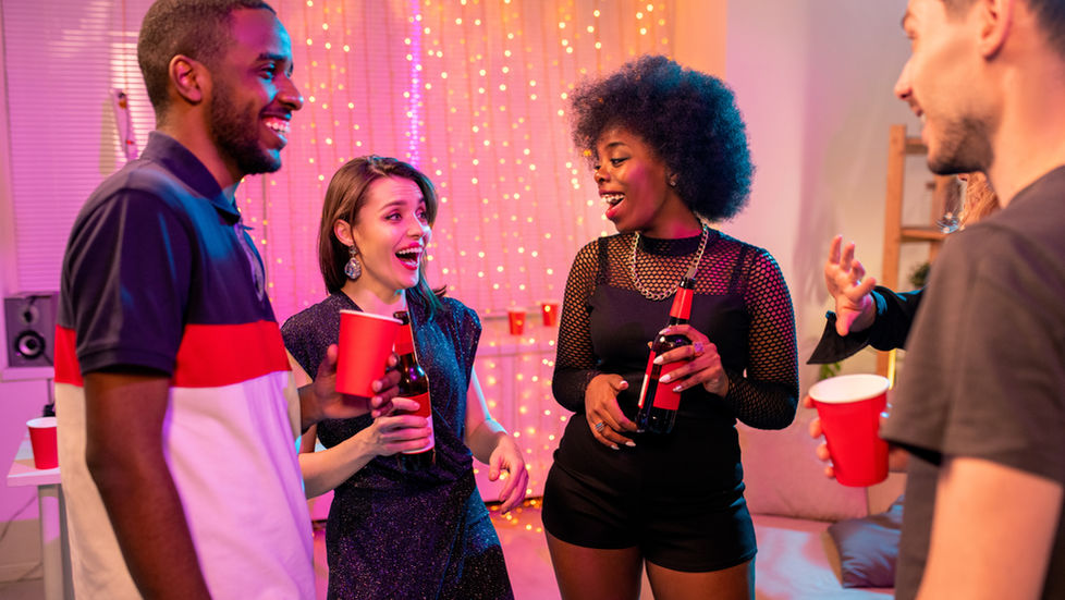 Two joyful young intercultural couples in glamorous attire having drinks at party in home environment and discussing funny stuff