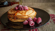 A stack of fluffy pancakes adorned with frozen raspberries, drizzled with rich syrup, elegantly presented on a dark plate against a burlap background.