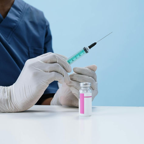 A healthcare professional wearing gloves and blue scrubs prepares a syringe with vaccine from a vial on a white table.