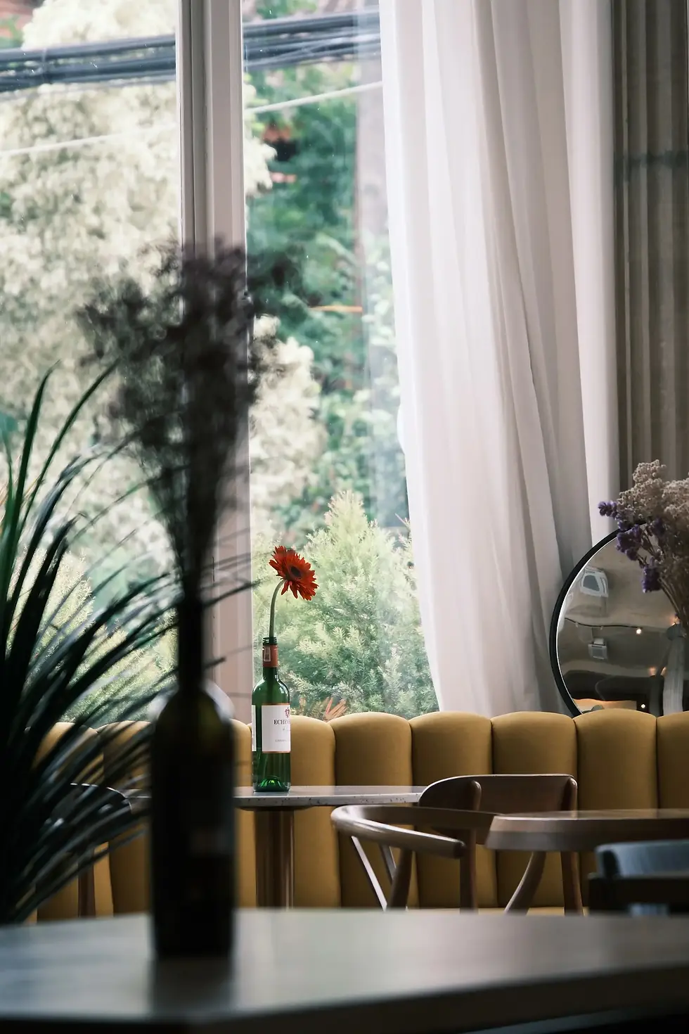 A cozy cafe with a yellow cushioned bench, a wine bottle with a red flower, wooden chairs, and large window showcasing greenery outside.