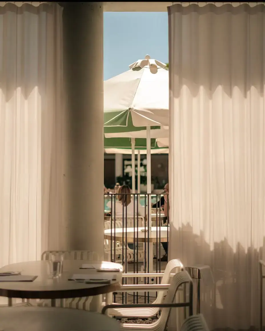 View through sheer curtains revealing a sunlit patio with a green and white umbrella and seated people. The scene is calm and inviting.