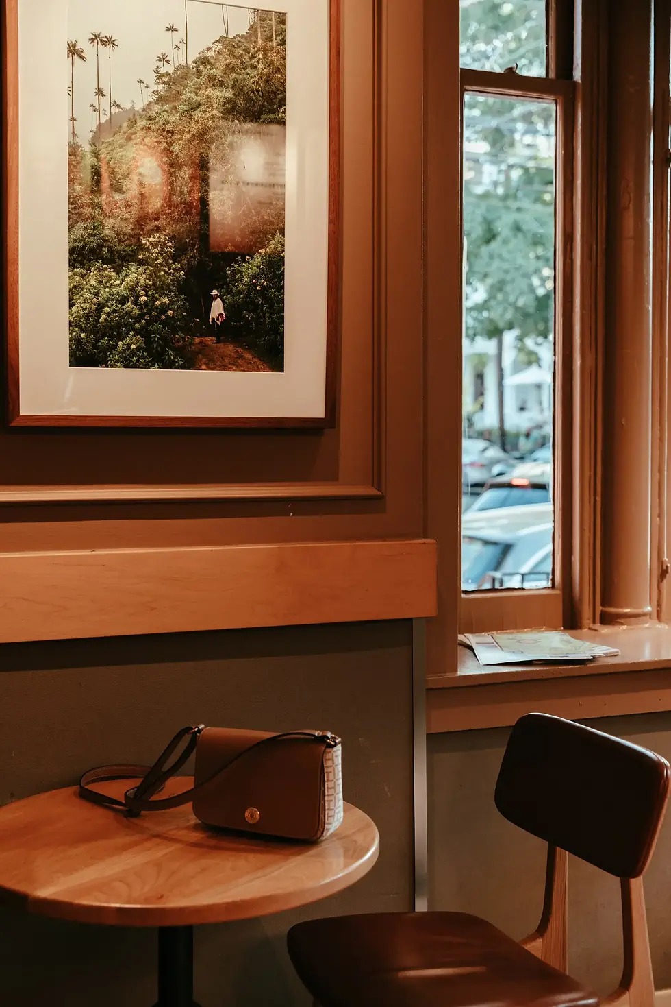 A cozy coffee shop corner with a brown purse on a round table. Framed landscape photo on the wall, window view of parked cars outside.
