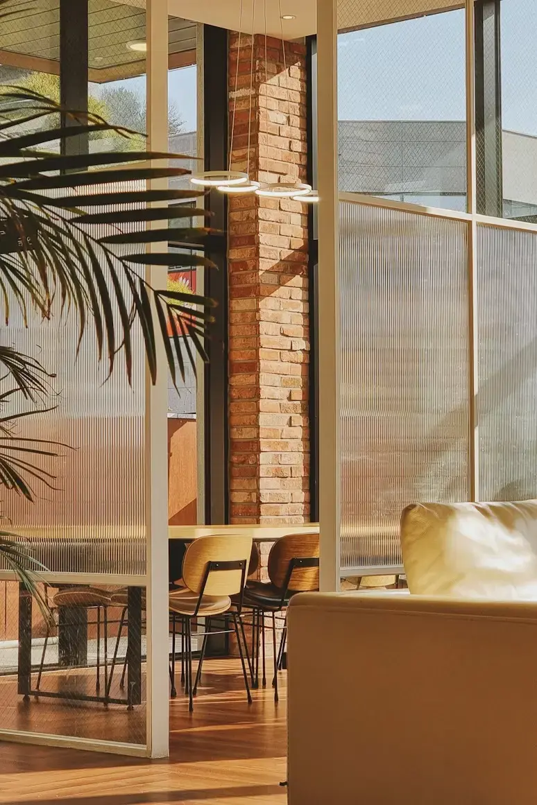 Cozy interior with a sunlit dining table, wood floor, and brick walls. A palm leaf peeks in, adding a touch of greenery. Peaceful ambiance.