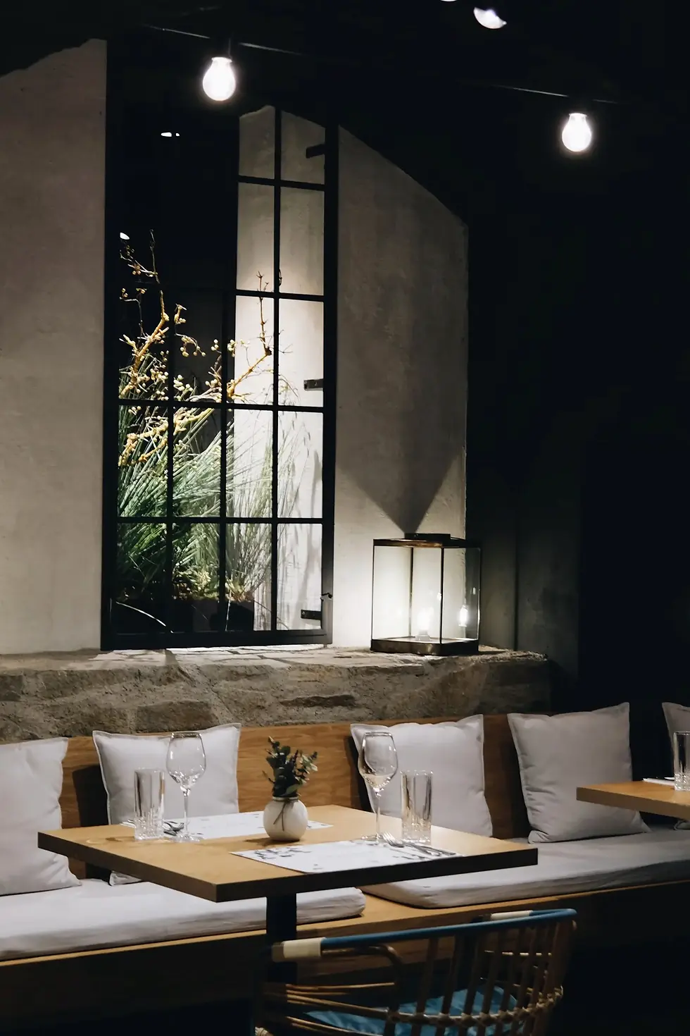 Stylish cafe interior with wooden tables set with glasses and white cushions. Plants visible through a large window. Warm, cozy ambiance.