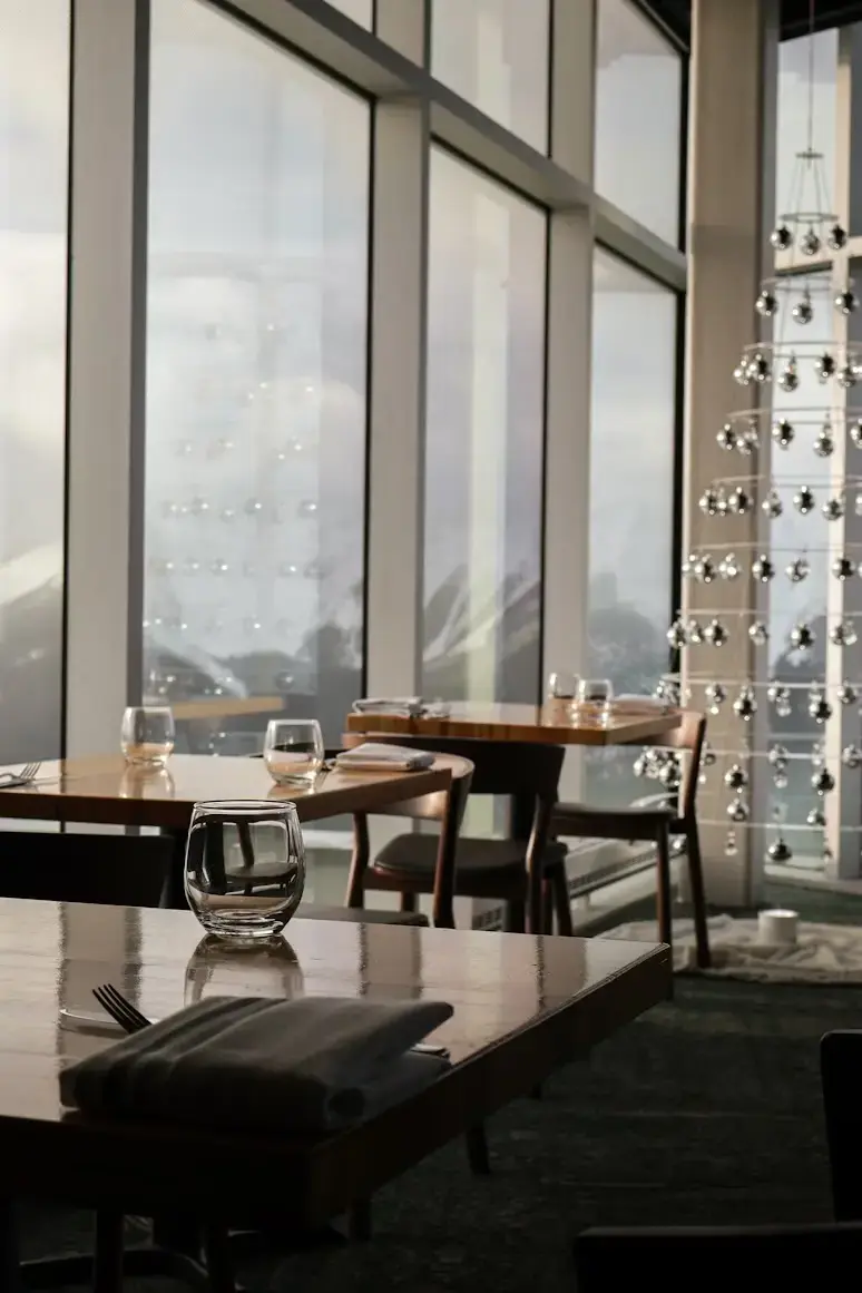 Modern restaurant with wooden tables, set with glassware and napkins, large windows showcasing a mountain view. Ambient and peaceful.