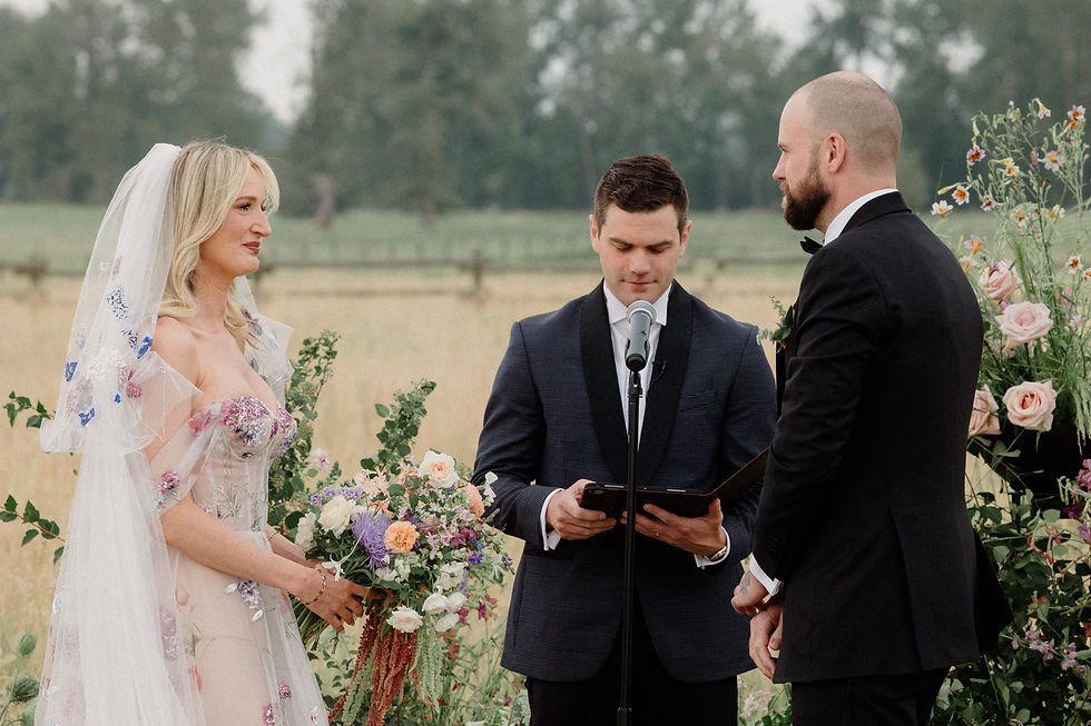 Bride and groom in floral attire exchange vows outdoors, surrounded by wildflowers. The officiant reads from a book, setting a serene mood.