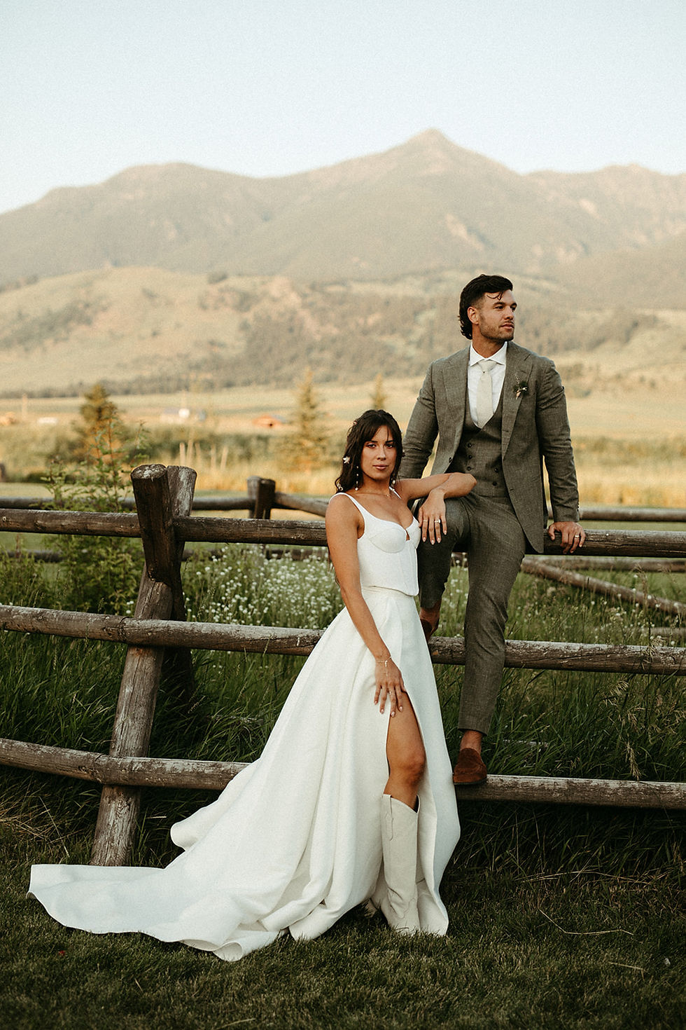 Bride in a white dress and groom in a gray suit pose by a wooden fence with scenic mountains in the background, exuding a serene vibe.