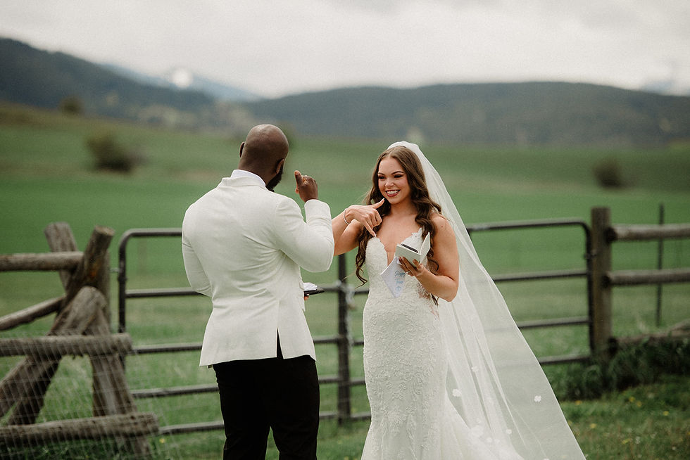 Bride in white gown reads vows to groom in white jacket, set in a lush green field with mountains. Both smiling, creating a joyful mood.