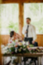 Man in tie giving speech with microphone, smiling woman in white dress seated, surrounded by colorful flowers, wood background, bright day.