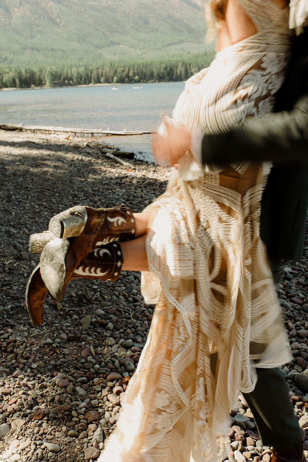 A person lifts someone in a lace dress and cowboy boots by a lake. Background shows trees, mountains, and a rocky shore. Mood is joyful.