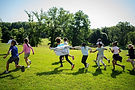 Children running in a grassy field on a sunny day with trees in background.