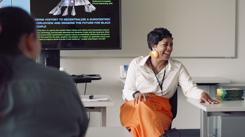 A smiling woman in a white shirt and orange skirt sits on a desk in a classroom, with a presentation screen and books nearby.