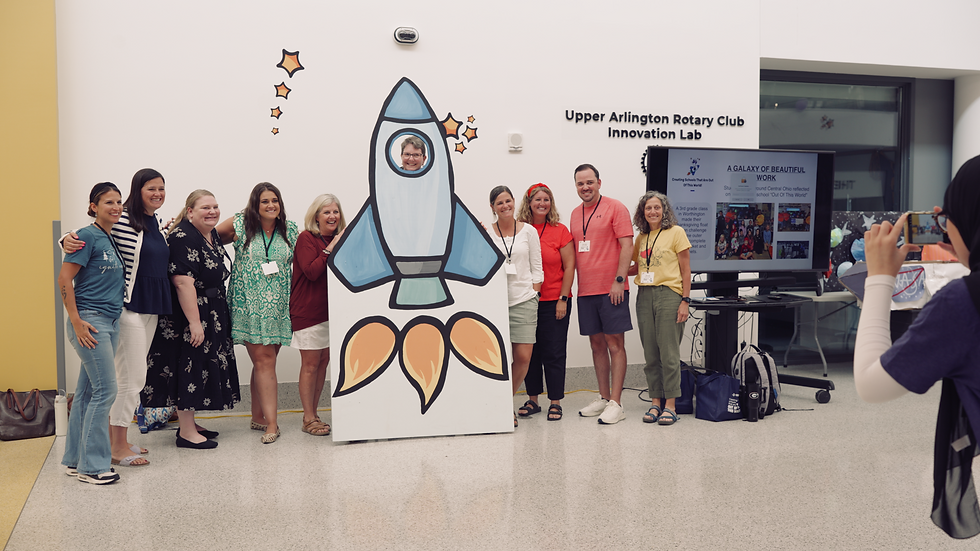 Group of people smiling by a rocket cutout in the Upper Arlington High School hallway. A person takes a photo, and a TV displays text.