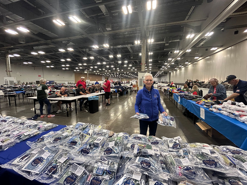 A woman in a blue jacket holds packaged items in a large convention hall with tables covered in clothing. Bright overhead lights illuminate the scene.