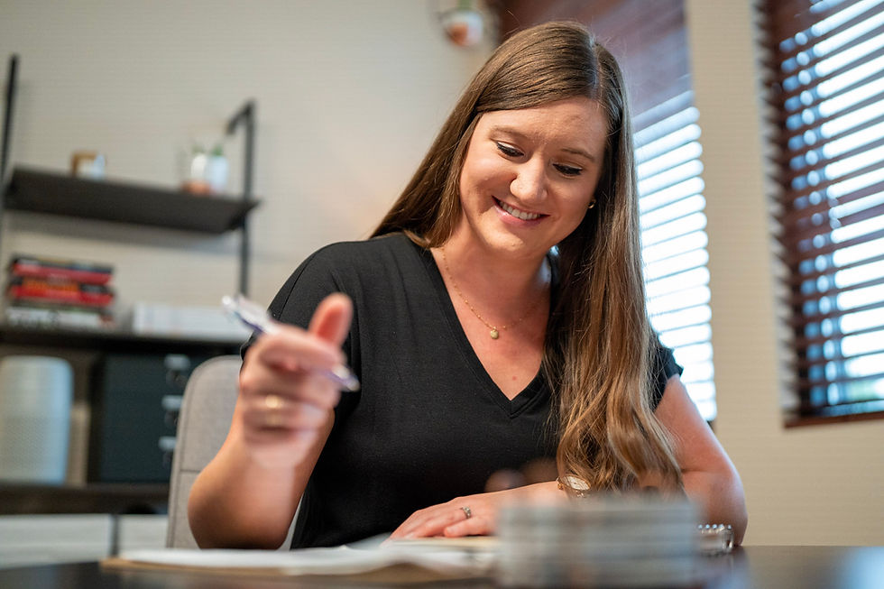 Woman in a black shirt writing at a desk, smiling. Blurred shelves and window blinds in the background. Bright and focused atmosphere.