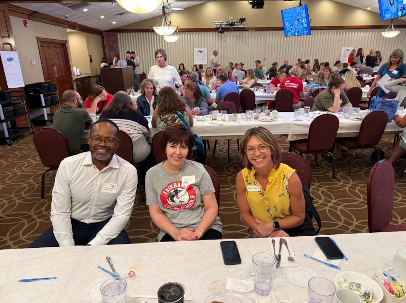 Three smiling people sit at a long table in a busy conference room. The woman in yellow wears glasses. Background shows crowded tables and screens.