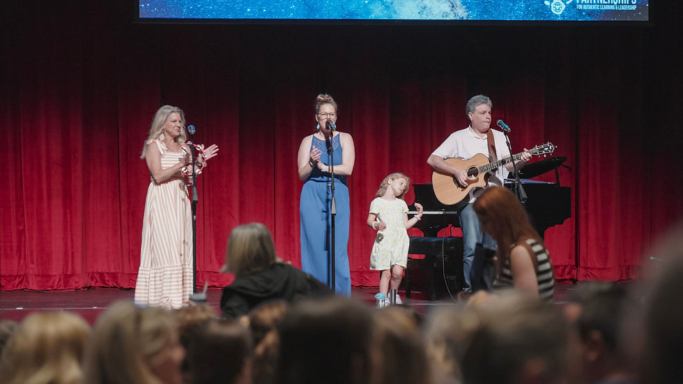 Four people performing on stage; one plays guitar, two sing and clap, a child stands nearby. Red curtains backdrop. Audience in foreground.