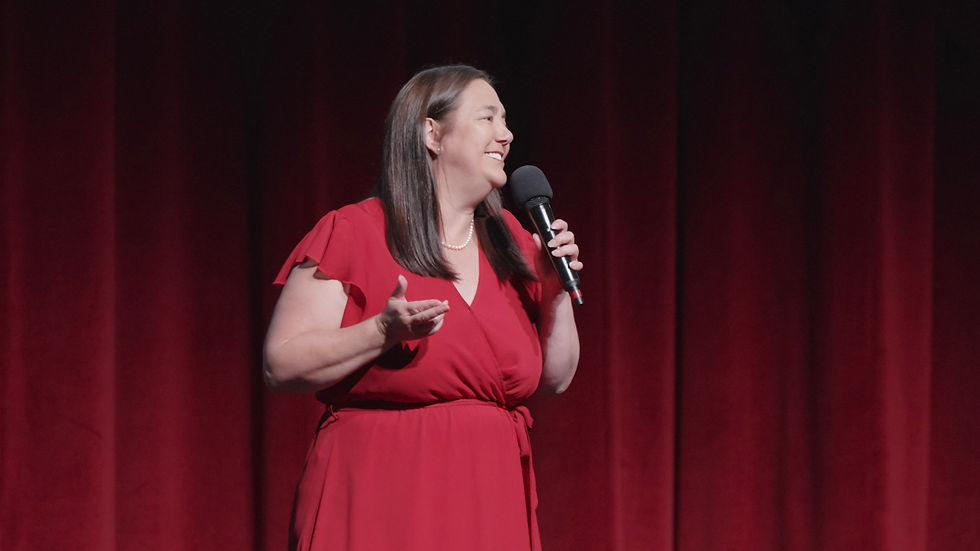Woman in a red dress speaking into a microphone on stage with red curtains in the background, smiling and gesturing with her hand.