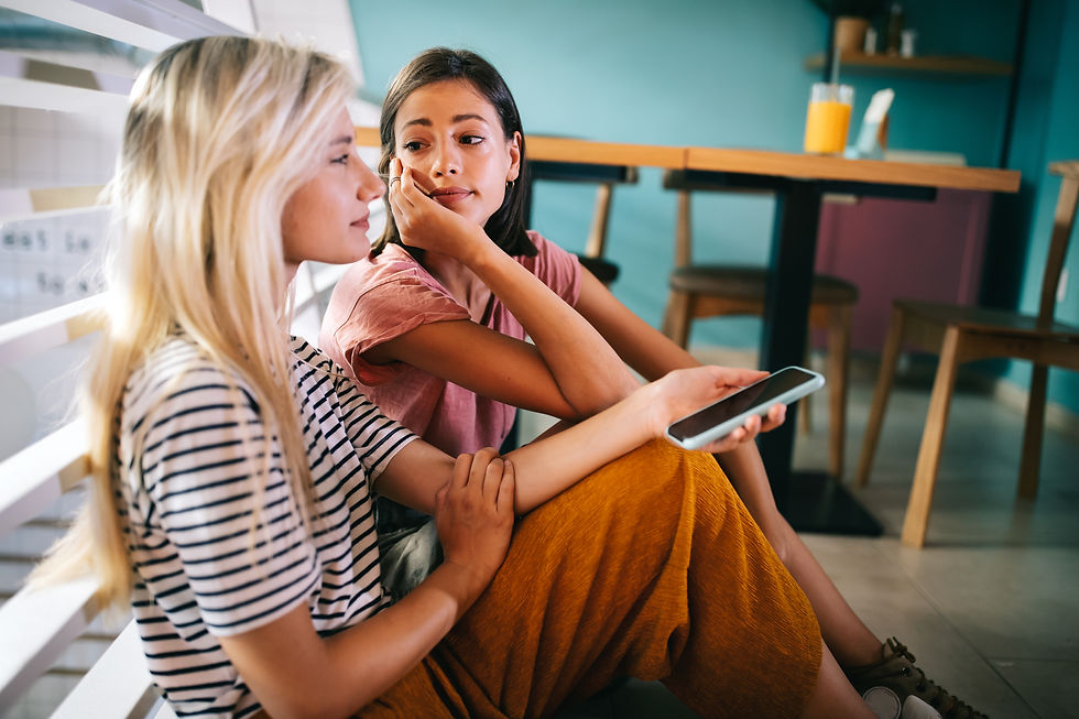 Two women sit on a cafe floor, one holds a smartphone, deep in conversation. Bright attire matches the colorful, relaxed setting.