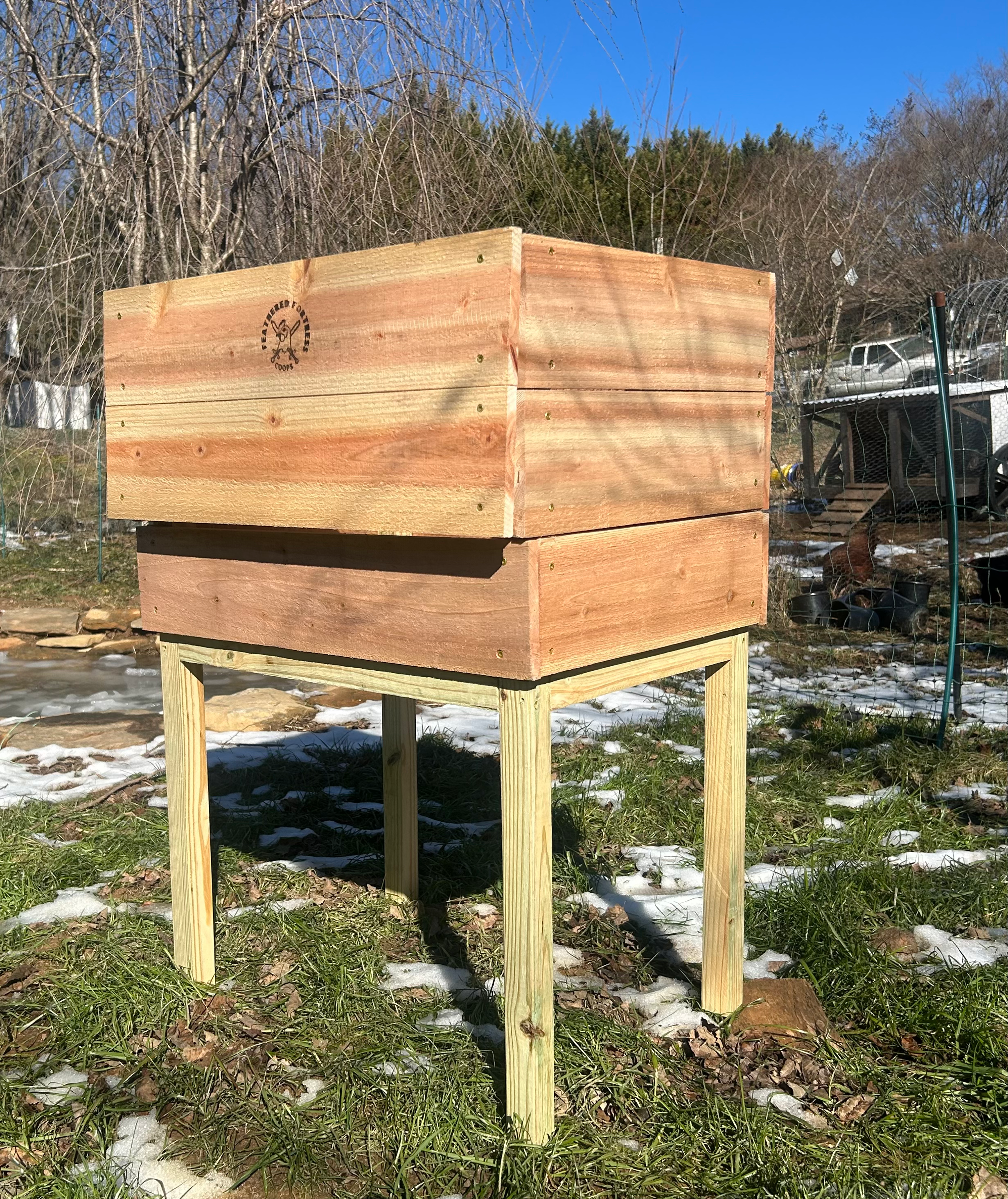 A chick brooder made from cedar in grass with patches of snow. 