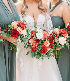 Bride and bridesmaids holding coordinated wedding bouquets with white, red, orange, and greenery accents.