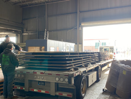 Flatbed truck being loaded inside a warehouse with large sheets of polished stainless steel, as workers stack and secure the metal plates for transport.