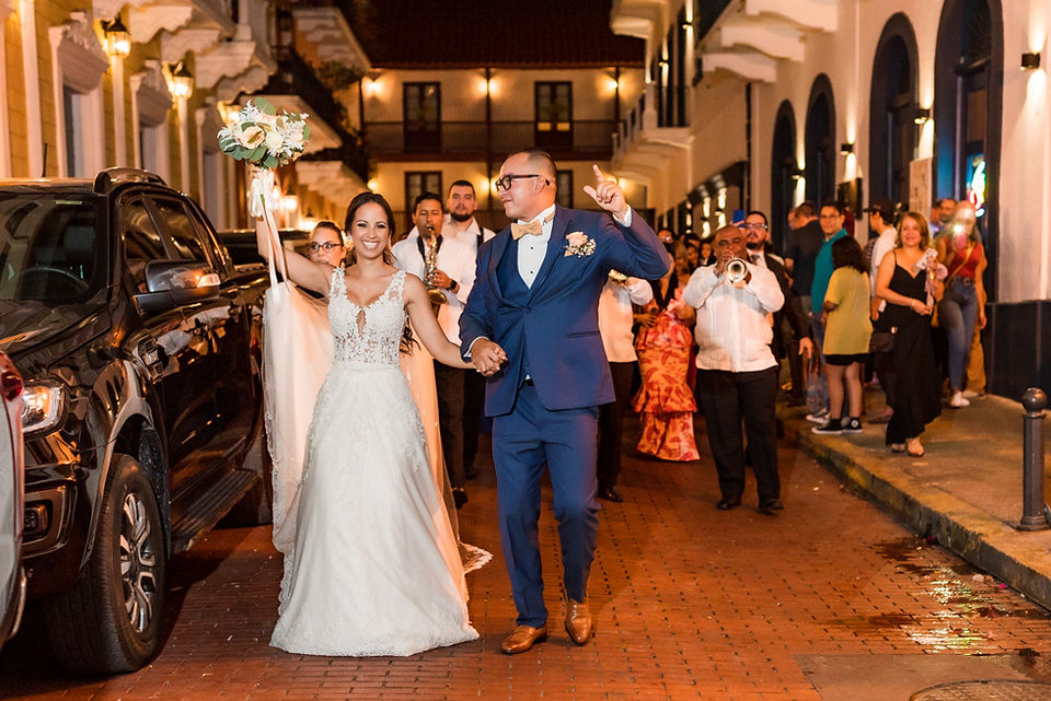 Novios recién casados caminando y bailando tomados de la mano por las calles del Casco Antiguo durante una tuna de carnaval; la novia alza su ramo con la mano derecha mientras el novio la mira fijamente y feliz, y al fondo se ven los músicos e invitados disfrutando y celebrando su boda. Ciudad de Panamá. Fotografía por Ahmed Saturno.