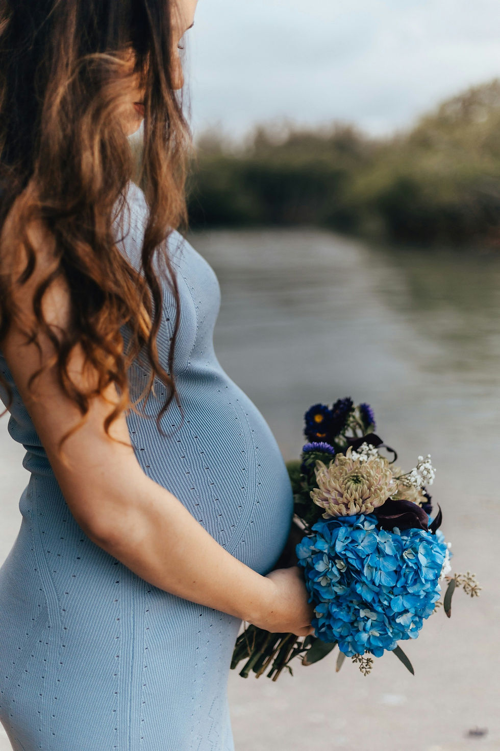 Pregnant woman in a blue dress holding a bouquet of blue hydrangeas and other flowers by a lake