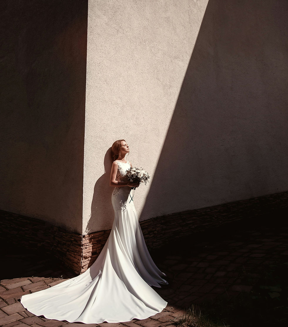 Bride in a white wedding dress holding a bouquet, standing in front of a white wall with sunlight