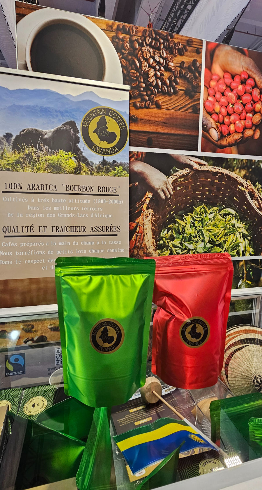 Stand Mountain Coffee Rwanda au Salon International de l'Agriculture de Paris, avec deux sachets de café debout (vert et rouge) portant le logo Mountain Coffee Rwanda, une affiche présentant le 100% Arabica Bourbon Rouge cultivé à 1800–2000m dans la région des Grands Lacs d'Afrique, le logo Fairtrade visible en bas à gauche, un petit drapeau rwandais et des objets artisanaux en arrière-plan.