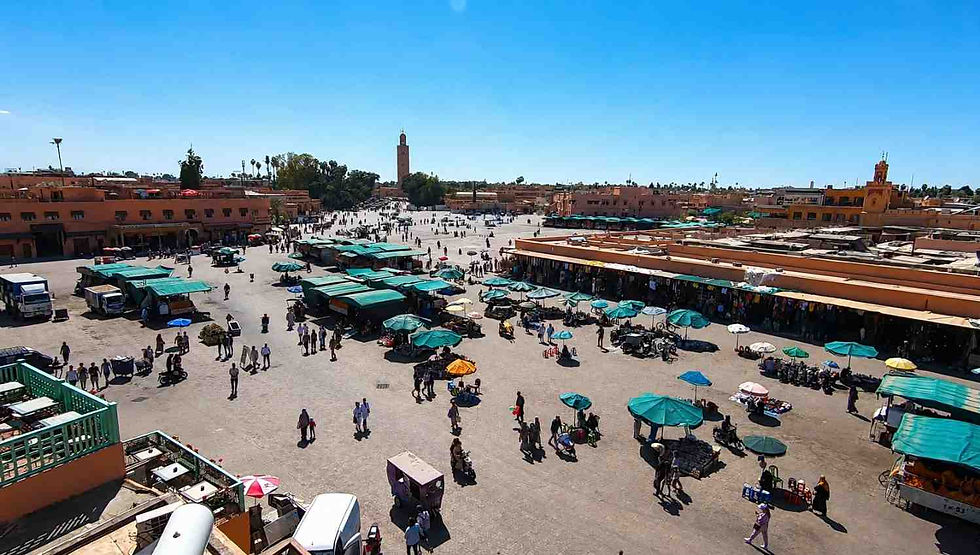 Midday view of Jemaa el‑Fnaa marketplace in Marrakesh, Morocco, captured from above under bright daylight. Green umbrellas and awnings shade vendor stalls across the square, surrounded by terracotta buildings and the Koutoubia Mosque’s minaret rising in the background. The scene shows a few people walking without visible faces, emphasizing the souk’s authentic architecture and vibrant atmosphere