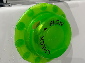 A bath overflow testing device positioned under the overflow opening of a bathtub, with a small bottle of water being poured into it to simulate a full bath test.
