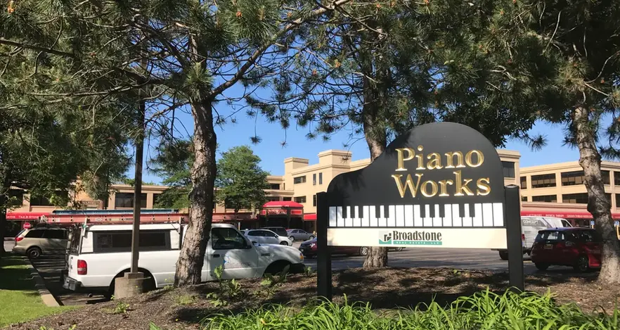 Piano Works sign with piano keys, near some cars and green bushes.