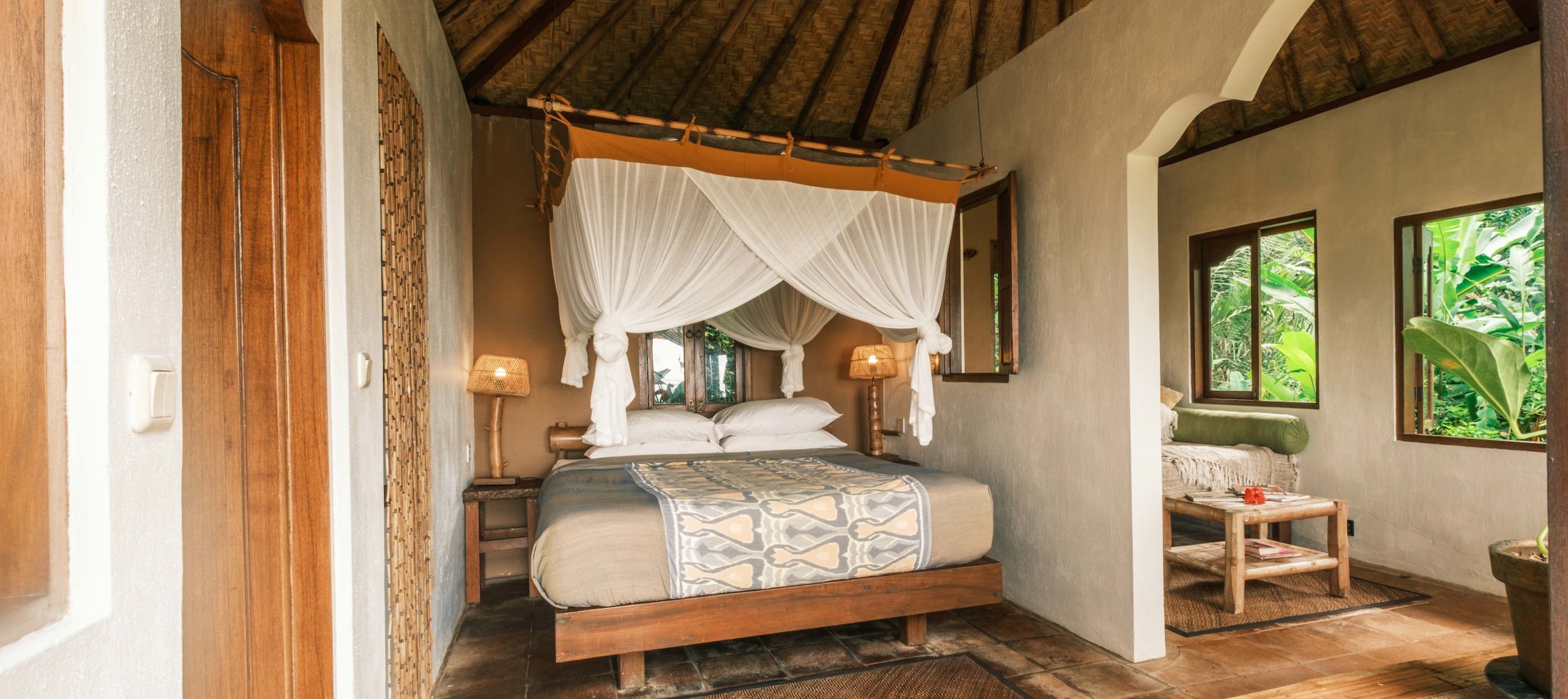 A bedroom with a four poster bed under a white net, timber floors and open windows onto the trees.