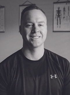 black and white photo of alex bishop, a smiling adult white man with short hair and a black shirt sits in the treatment room